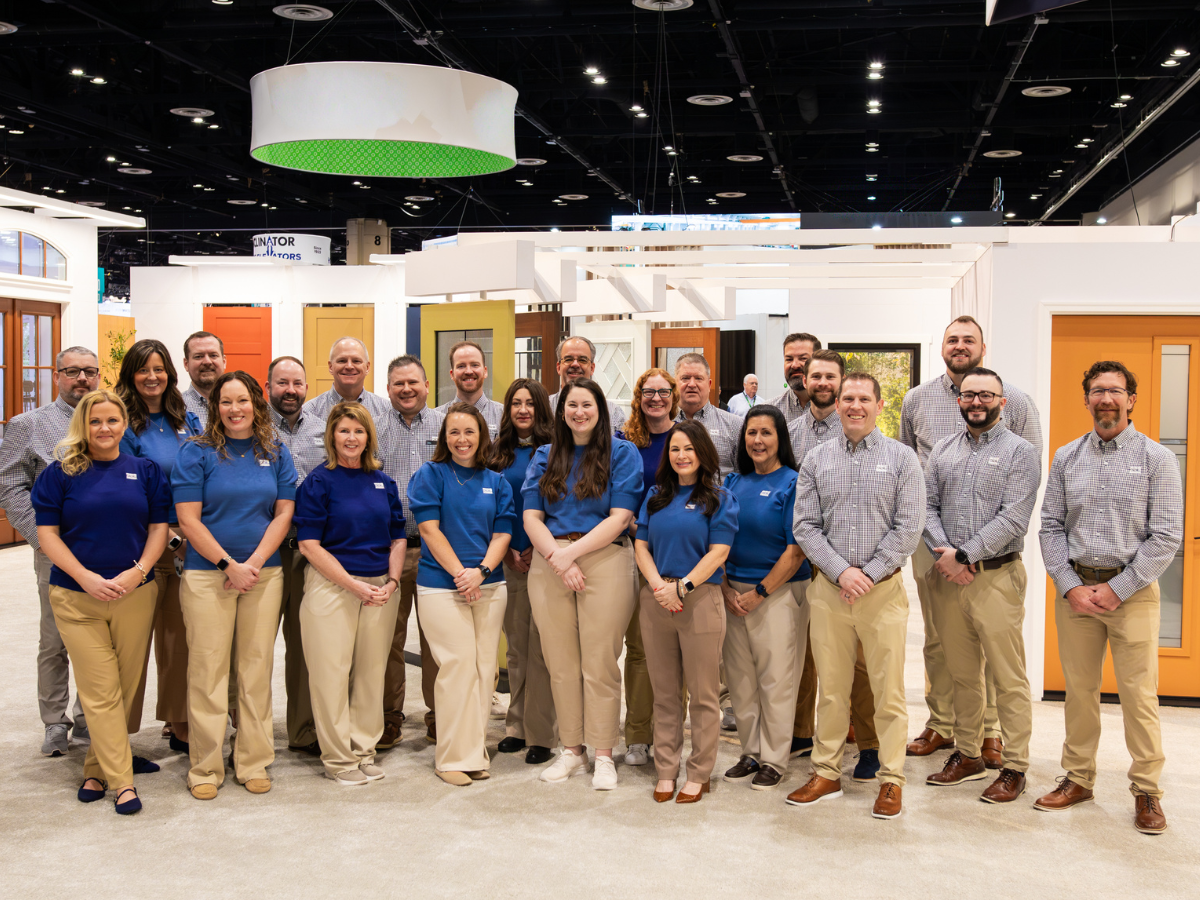 A large group of men and women posing for a photo in front of various door displays at a trade show, wearing blue shirts or light plaid shirts.